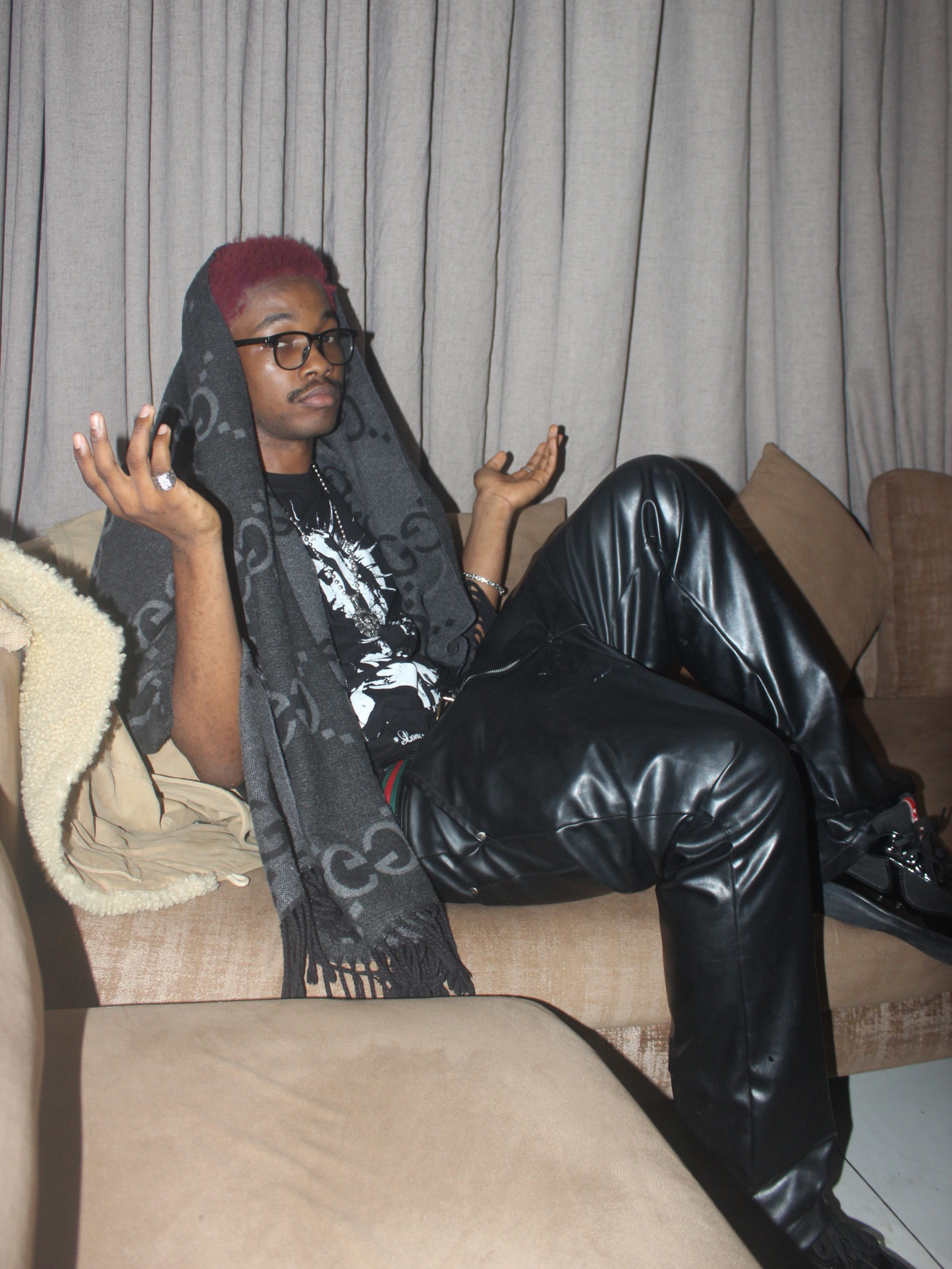 A person with short red hair and glasses lounges on a beige sofa, wearing a black graphic T-shirt, oversized black leather trousers and a long dark scarf draped over their head, with both hands raised in a relaxed pose.