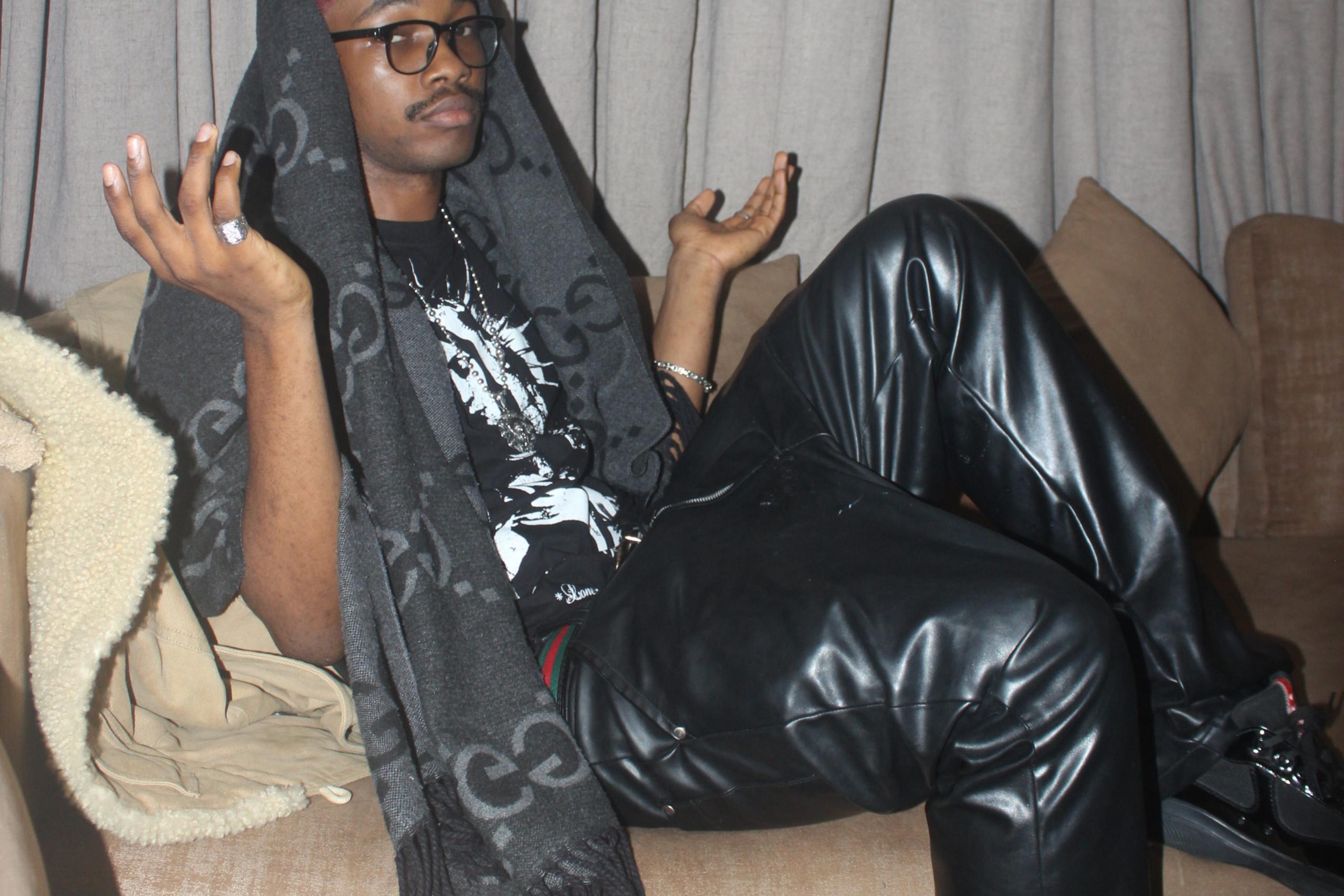 A person with short red hair and glasses lounges on a beige sofa, wearing a black graphic T-shirt, oversized black leather trousers and a long dark scarf draped over their head, with both hands raised in a relaxed pose.