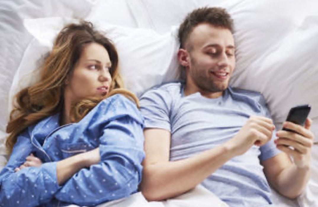 couple in bed, she is suspiciously looking at him using his phone