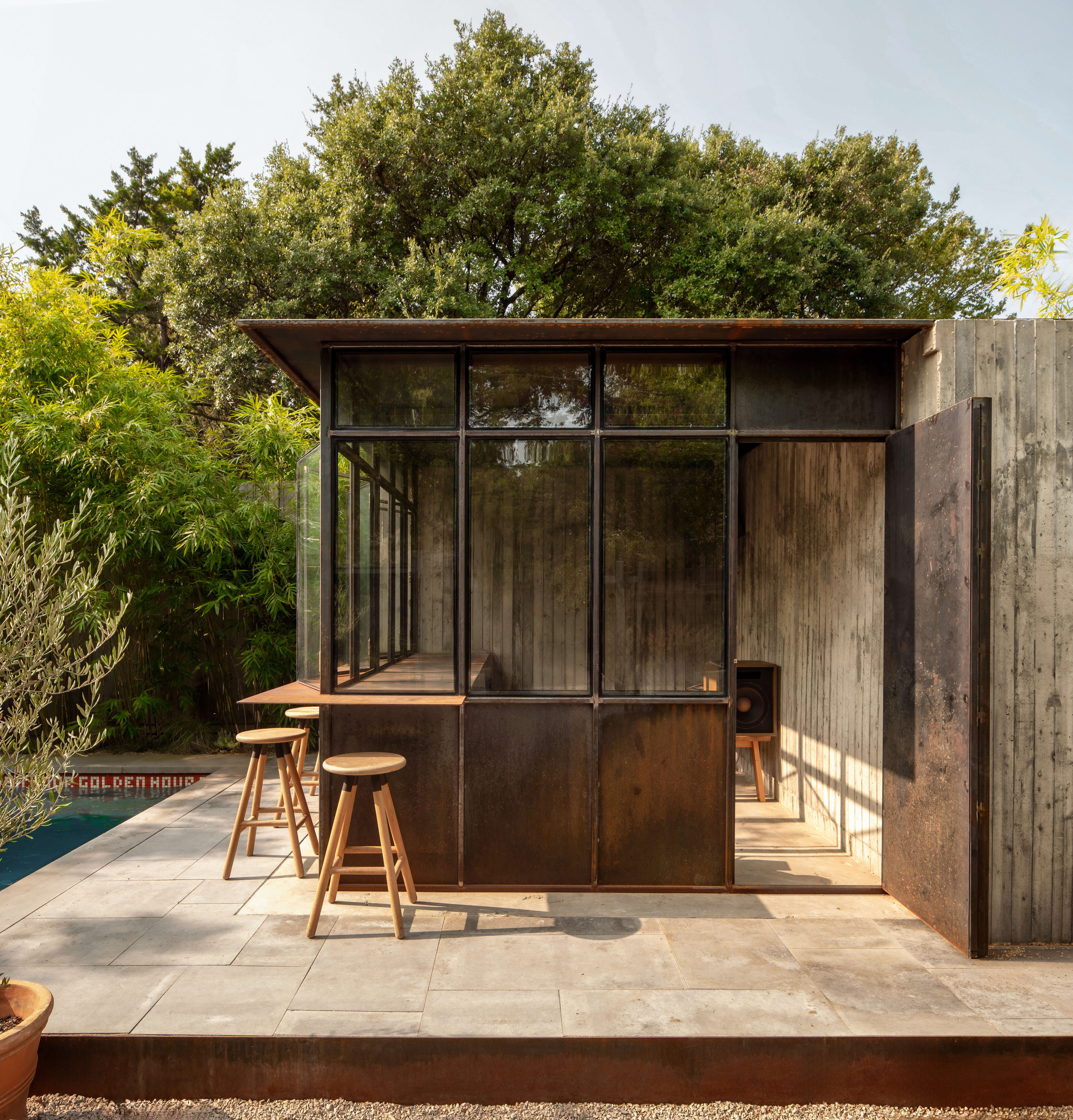 Corten steel pool house exterior with glass bar wall, surrounded by mature Dallas trees