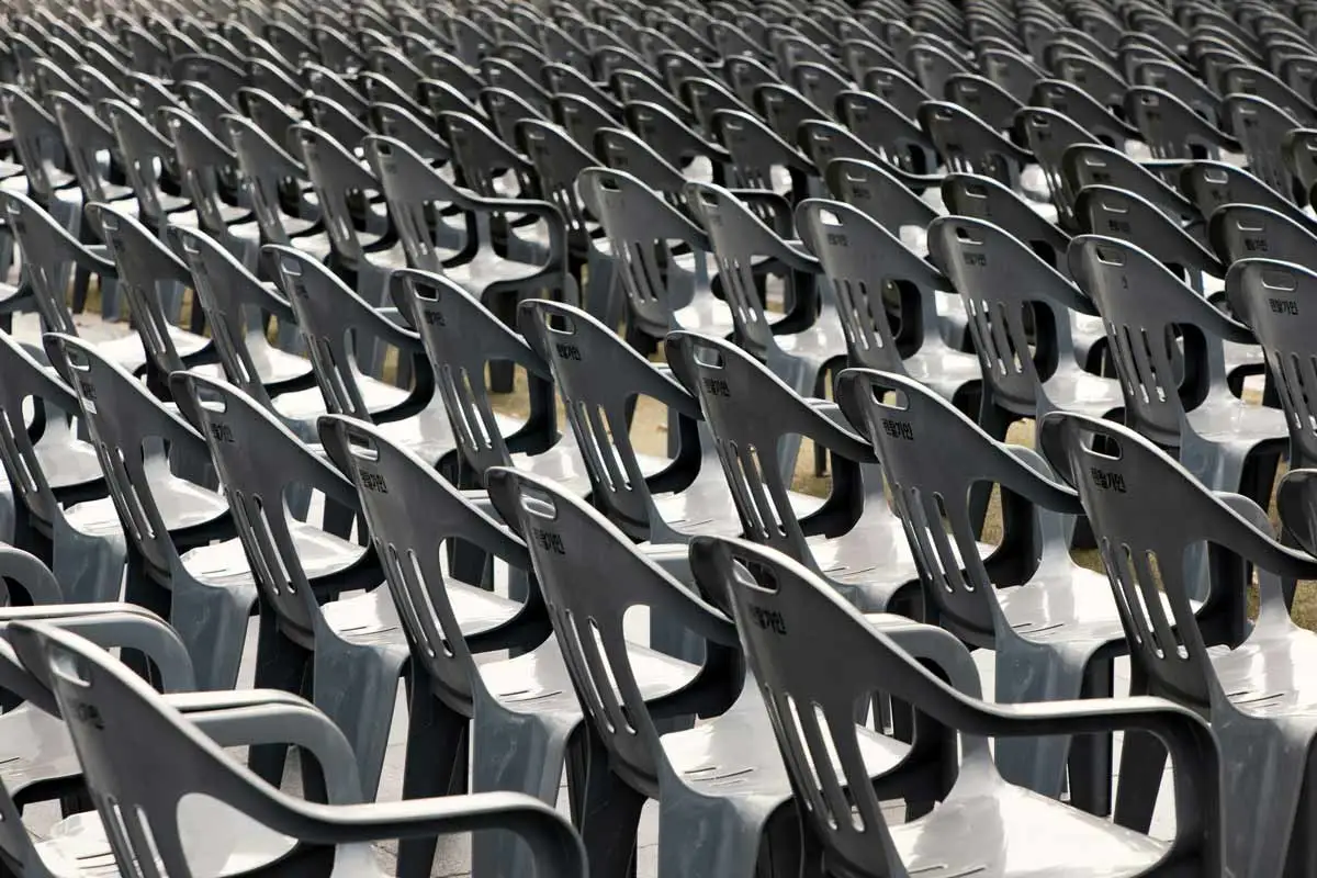 A photograph showing hundreds of identical gray plastic stackable chairs arranged in perfectly aligned, tight rows stretching into the distance. The perspective is from a low angle, emphasizing the repetition and scale of the seating. The chairs are empty, creating a sterile and uniform pattern that dominates the entire frame.
