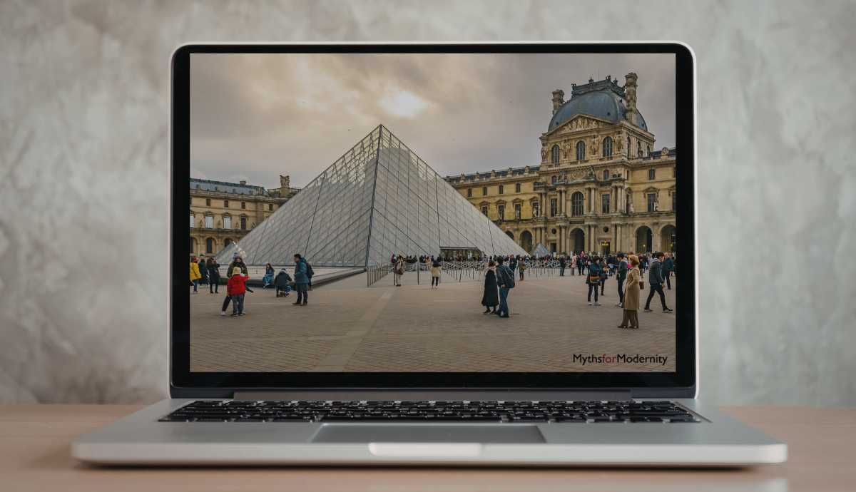 A silver laptop on a wooden desk displaying a high-resolution photograph of the Louvre Museum's glass pyramid and historic palace, illustrating the digital interface of modern cultural institutions.