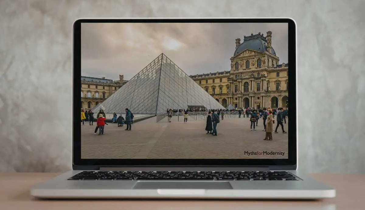 A silver laptop on a wooden desk displaying a high-resolution photograph of the Louvre Museum's glass pyramid and historic palace, illustrating the digital interface of modern cultural institutions.