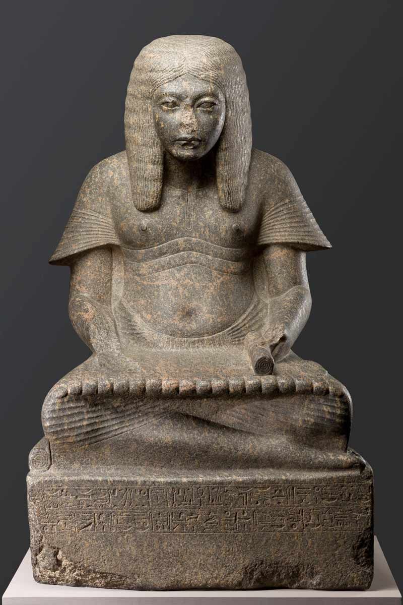A front-facing view of an ancient Egyptian stone statue of a seated scribe. The figure has a shoulder-length wig and is cross-legged with a papyrus scroll resting on his lap. The base of the statue features detailed rows of carved hieroglyphics.