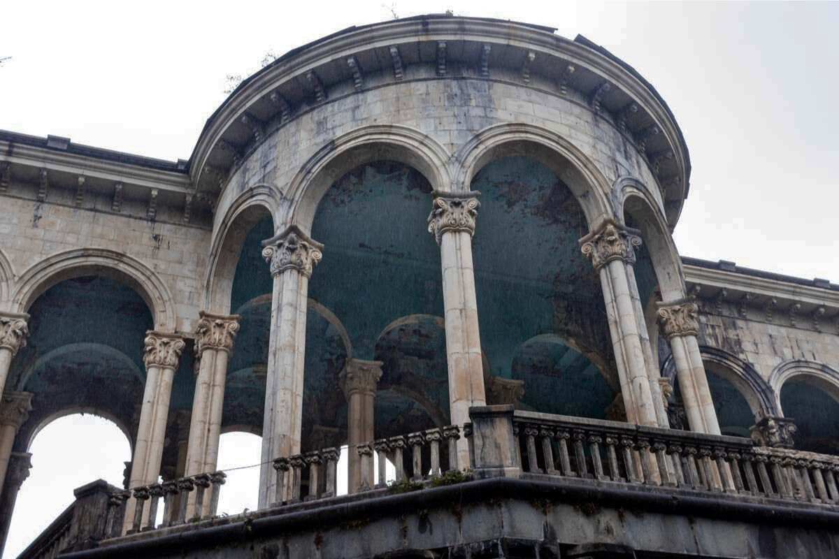 A low-angle shot of the iconic semi-circular balcony at Sanatorium Medea in Tskaltubo. Weathered stone columns support a series of elegant arches. The vaulted ceiling beneath is covered in vibrant but decaying blue paint that is peeling away to reveal the masonry. A stone balustrade lines the edge of the balcony against a bright, overcast sky.