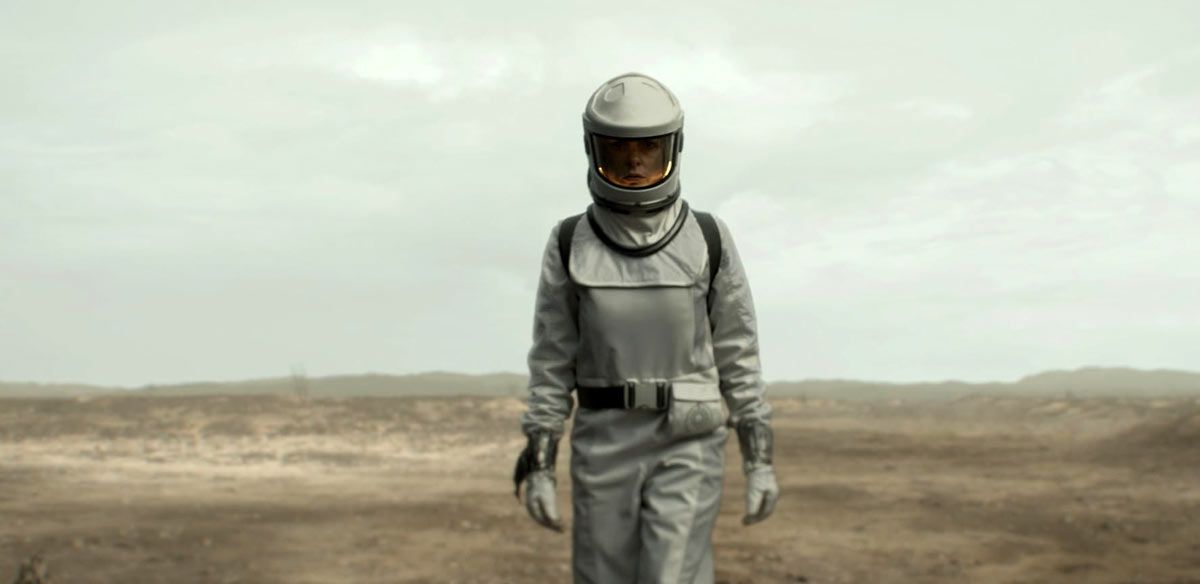 Juliette Nichols from the series Silo wearing a bulky, white protective hazmat suit and helmet with a reflective visor, walking toward the camera across a desolate, hazy, gray wasteland.