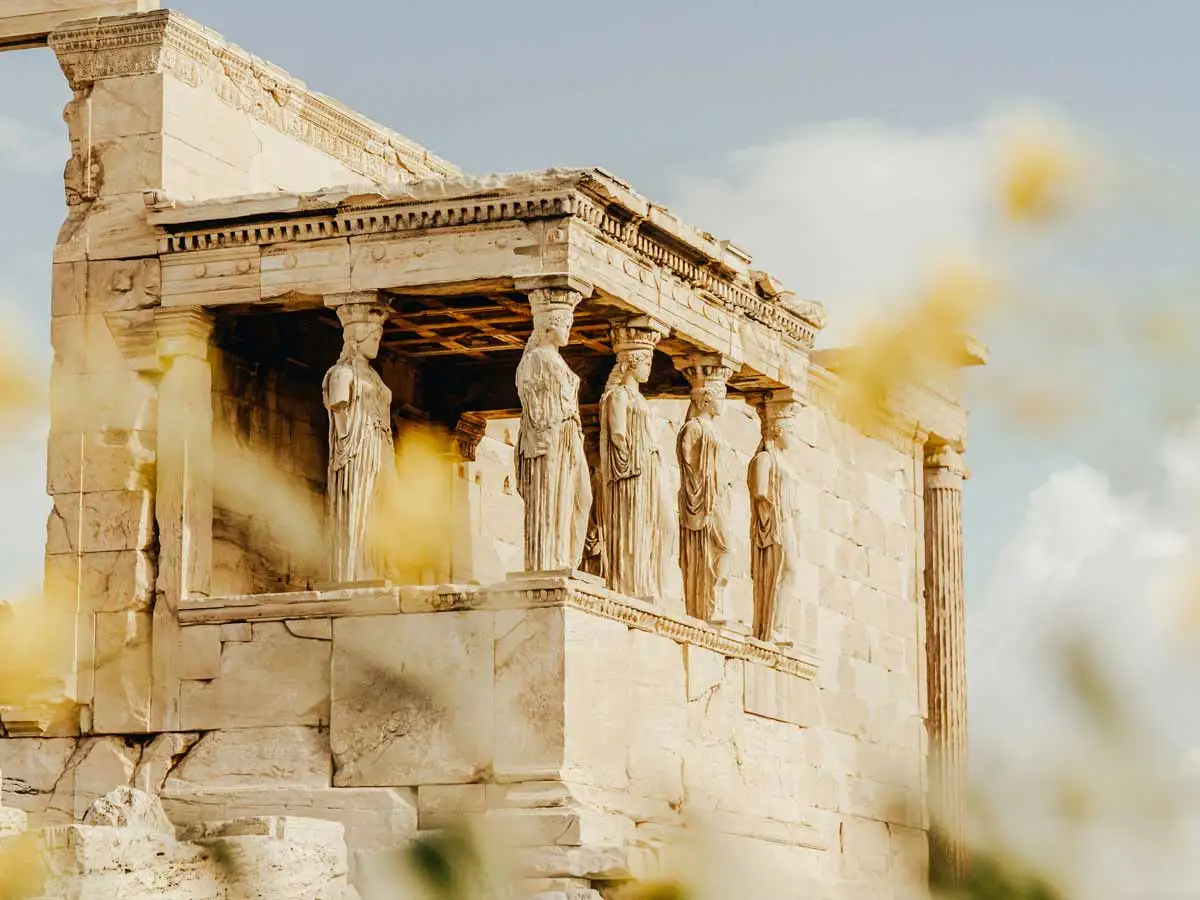 A high-angle, side-profile photograph of the Porch of the Maidens at the Erechtheion on the Acropolis of Athens. Six draped female figures, known as Caryatids, serve as architectural columns supporting the heavy stone roof of the porch. The ancient marble structure is bathed in warm, natural light against a pale sky, with soft-focus yellow flowers or foliage blurred in the foreground, creating a dreamy, atmospheric frame.