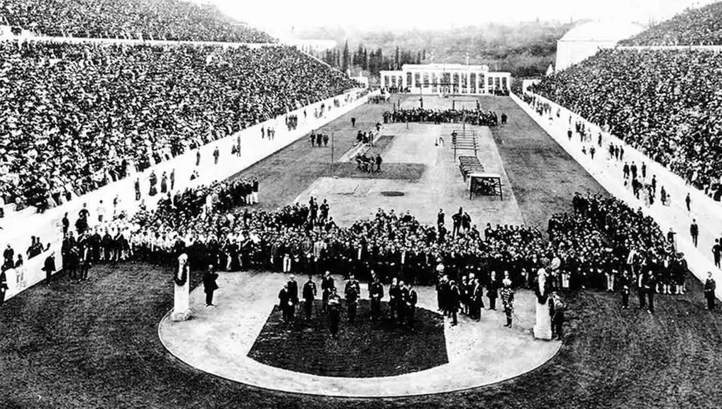 A historical black and white wide-angle photograph of a massive, densely packed U-shaped stadium, featuring rows of athletes and officials assembled on the central field during a formal ceremony.