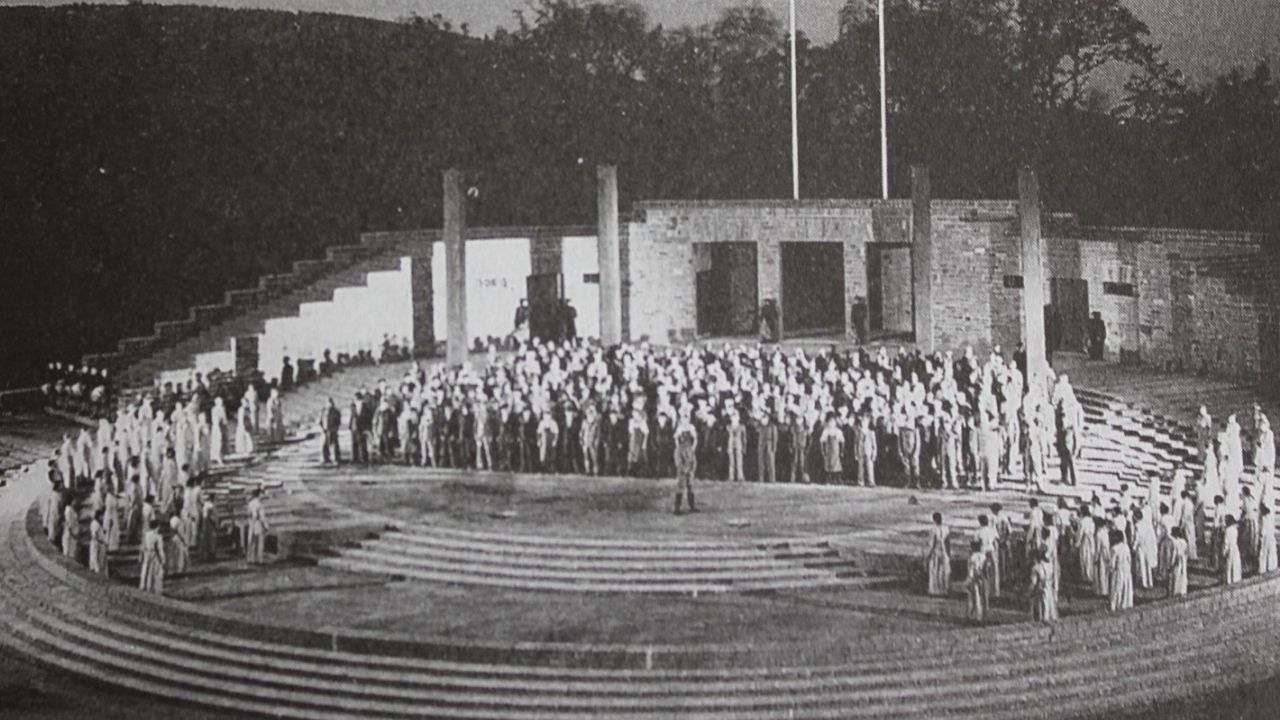 Black and white historical photo showing a large crowd of people standing on the curved stone steps and central stage of an outdoor, classical-style arena with tall pillars in the background.