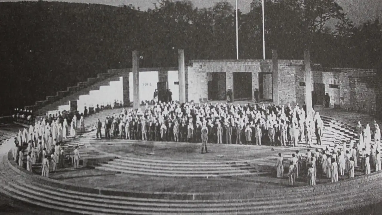 Black and white historical photo showing a large crowd of people standing on the curved stone steps and central stage of an outdoor, classical-style arena with tall pillars in the background.