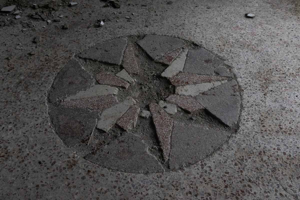 A top-down close-up of a cracked and damaged stone star inlay on a terrazzo floor. The circular mosaic is missing several pieces and is surrounded by dust and small fragments of stone.