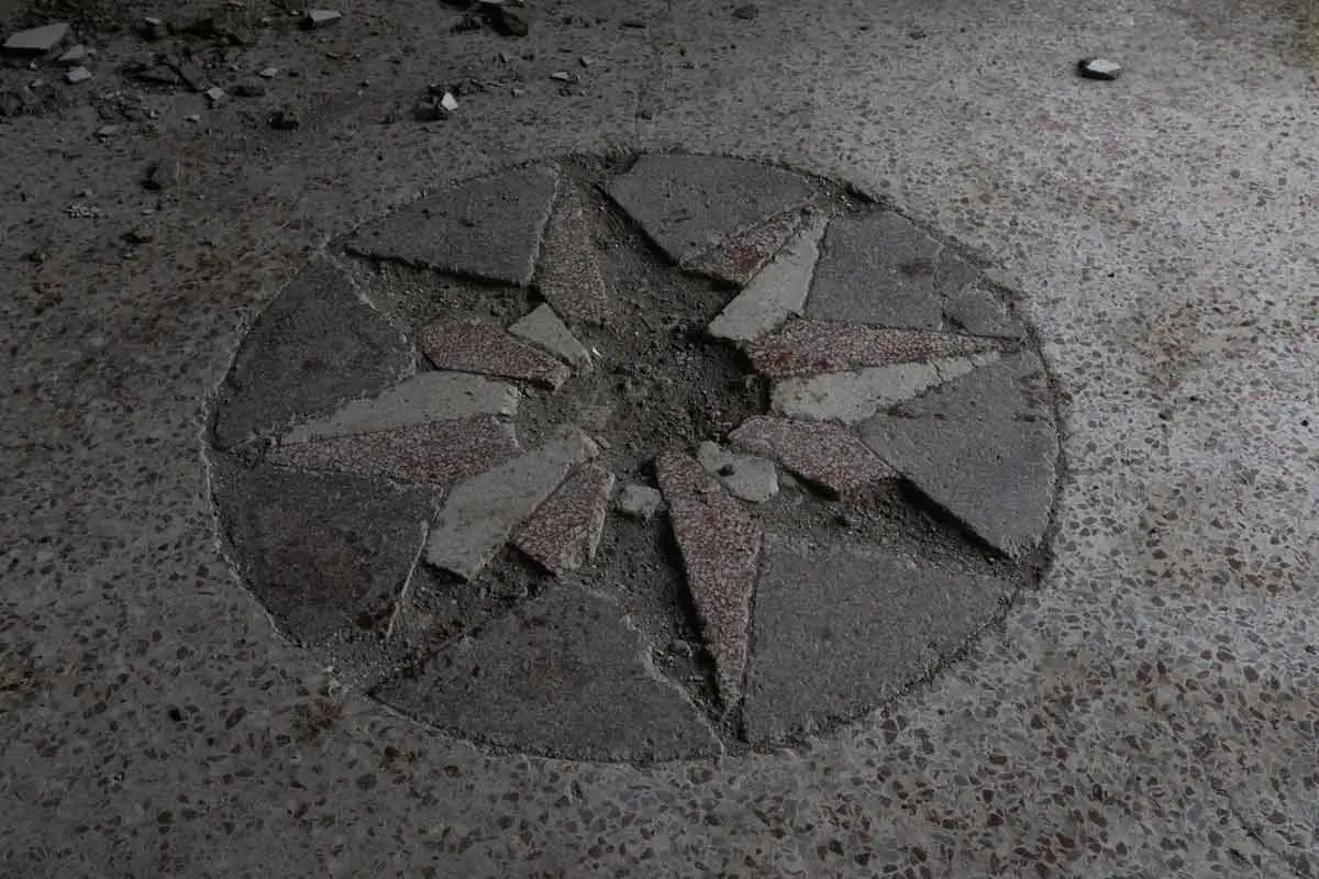 A top-down close-up of a cracked and damaged stone star inlay on a terrazzo floor. The circular mosaic is missing several pieces and is surrounded by dust and small fragments of stone.