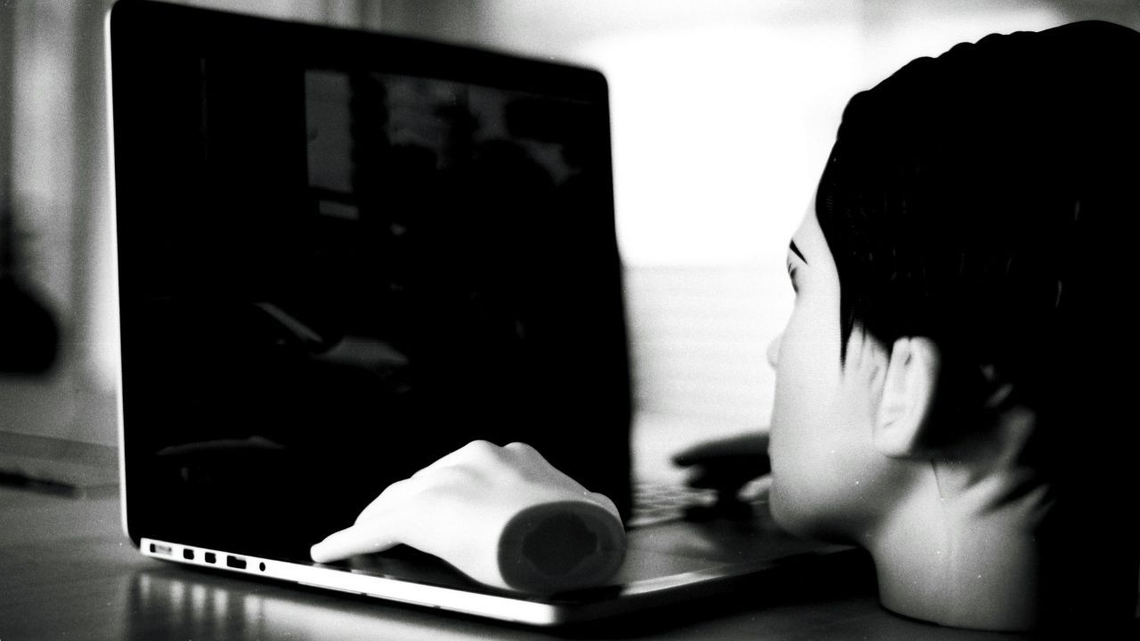 A black and white photograph showing a detached mannequin head and a disembodied mannequin hand positioned in front of an open laptop. The arrangement creates an eerie illusion of the lifeless mannequin staring into the dark screen and using the computer.