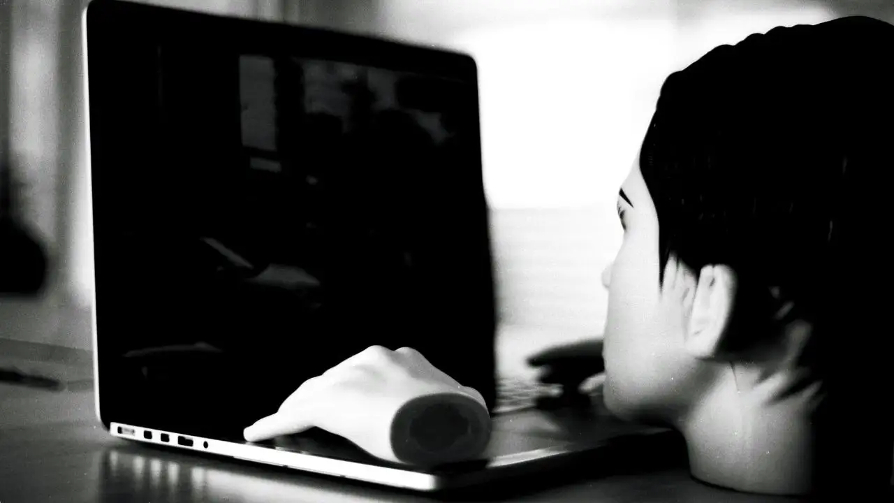 A black and white photograph showing a detached mannequin head and a disembodied mannequin hand positioned in front of an open laptop. The arrangement creates an eerie illusion of the lifeless mannequin staring into the dark screen and using the computer.