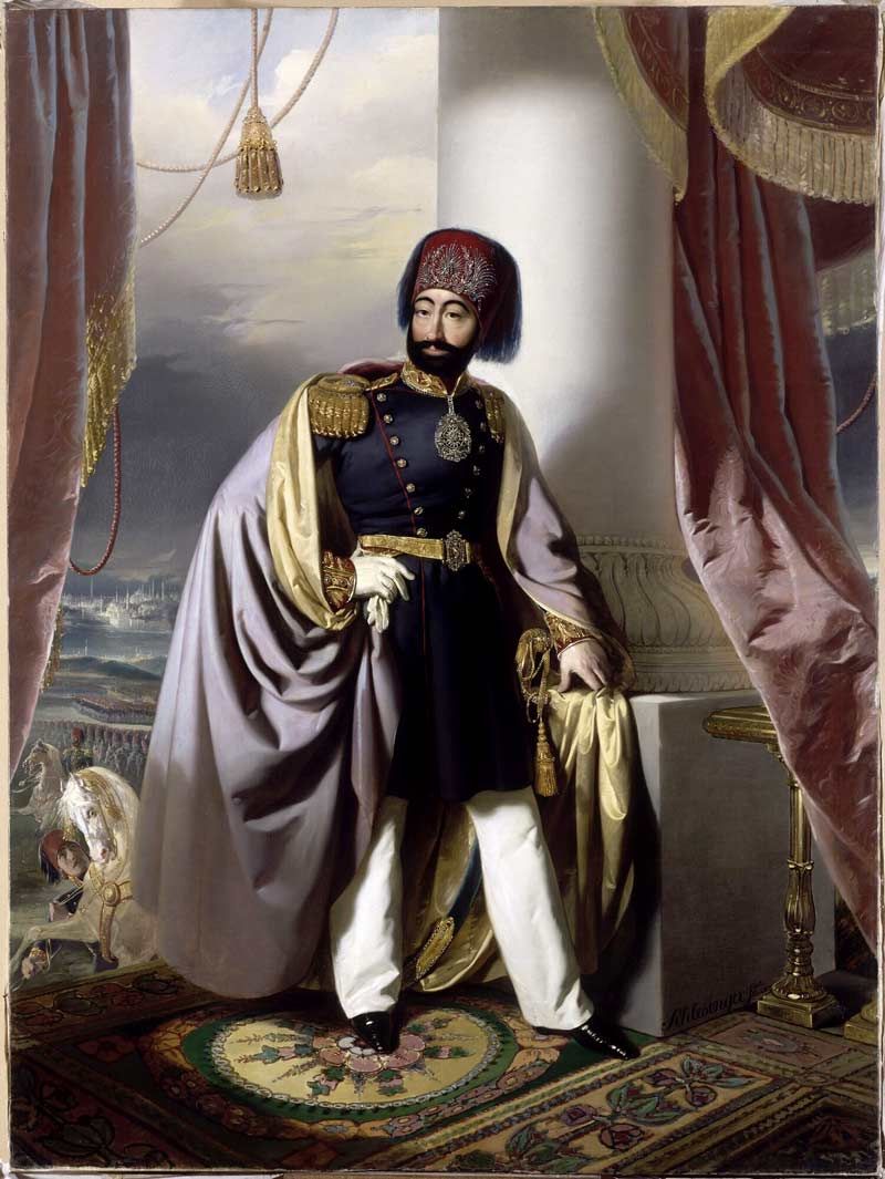 Henri Guillaume Schlesinger's full-length portrait painting of Sultan Mahmud II standing in a palace interior, wearing a decorated red fez, dark military uniform with gold epaulets, and a sweeping cape.