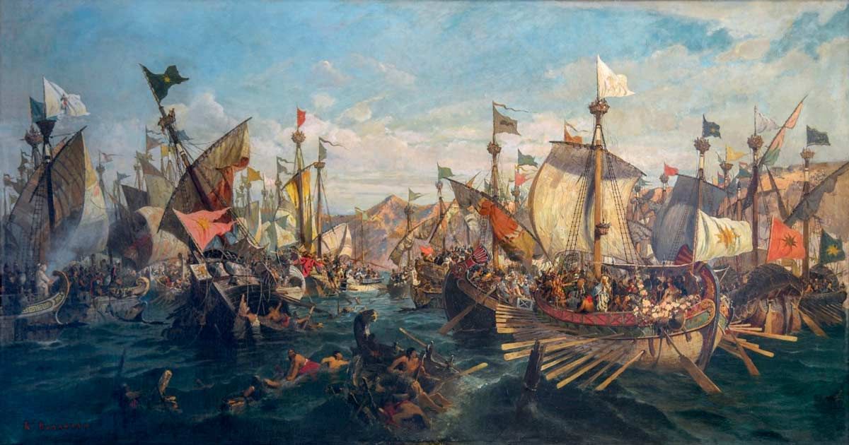 A dramatic oil painting by Konstantinos Volanakis depicting the naval Battle of Salamis. Numerous ancient triremes with large sails and multiple banks of oars are locked in a chaotic struggle on choppy, dark green water. The ships fly various colorful banners and flags. In the foreground, wreckage and soldiers are seen in the water, while distant brown hills sit under a cloudy sky.