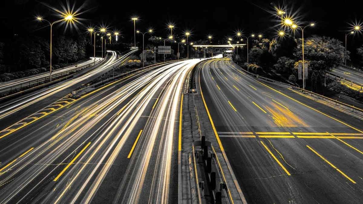 A long-exposure photograph of a busy multi-lane highway at night. The movement of traffic creates continuous, sweeping streaks of bright white and yellow light stretching into the distance under the glow of overhead streetlights, which flare into starburst patterns.