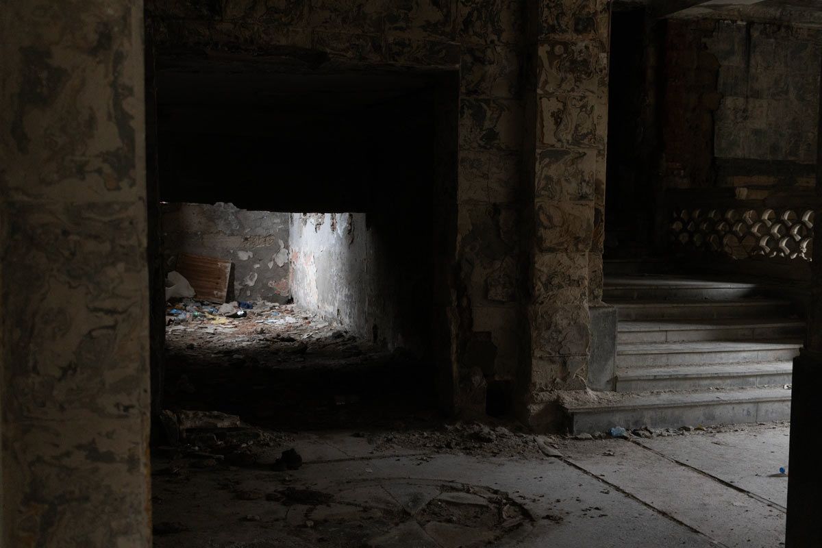 A dark, atmospheric interior of a crumbling stone building. A shaft of light from a side opening illuminates a floor covered in litter and debris, while a stone staircase leads upward into the darkness on the right.