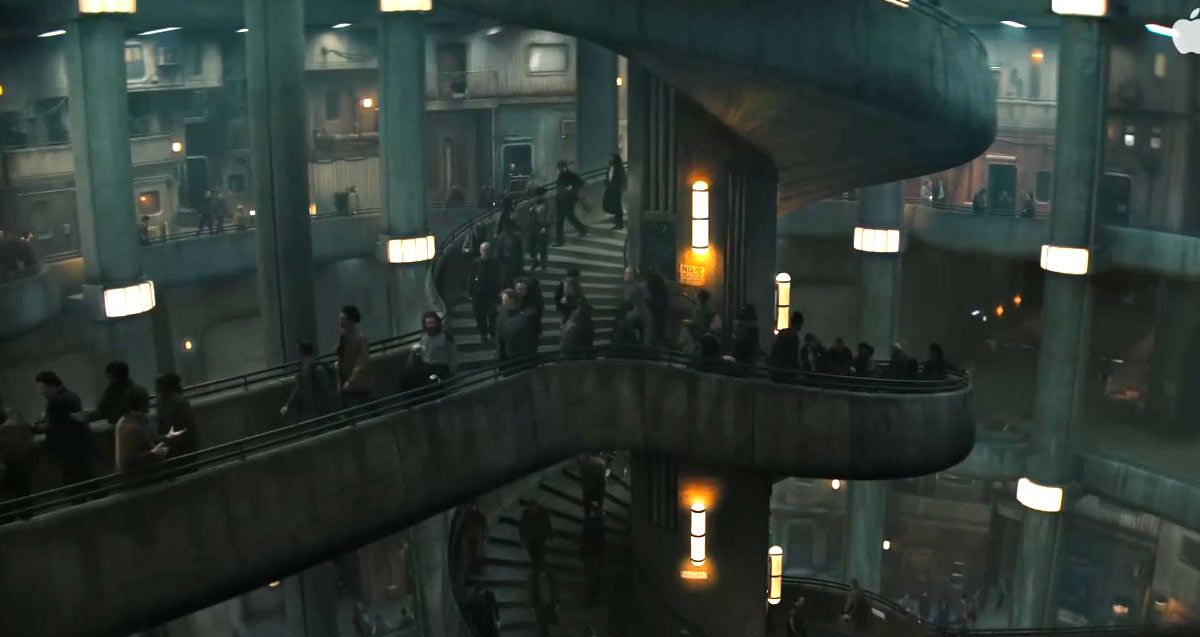 A wide, atmospheric shot of the vast, brutalist interior of the silo. A massive concrete spiral staircase winds down the center, connecting multiple levels of balconies and walkways where small figures of residents are seen walking under warm, artificial yellow lighting.