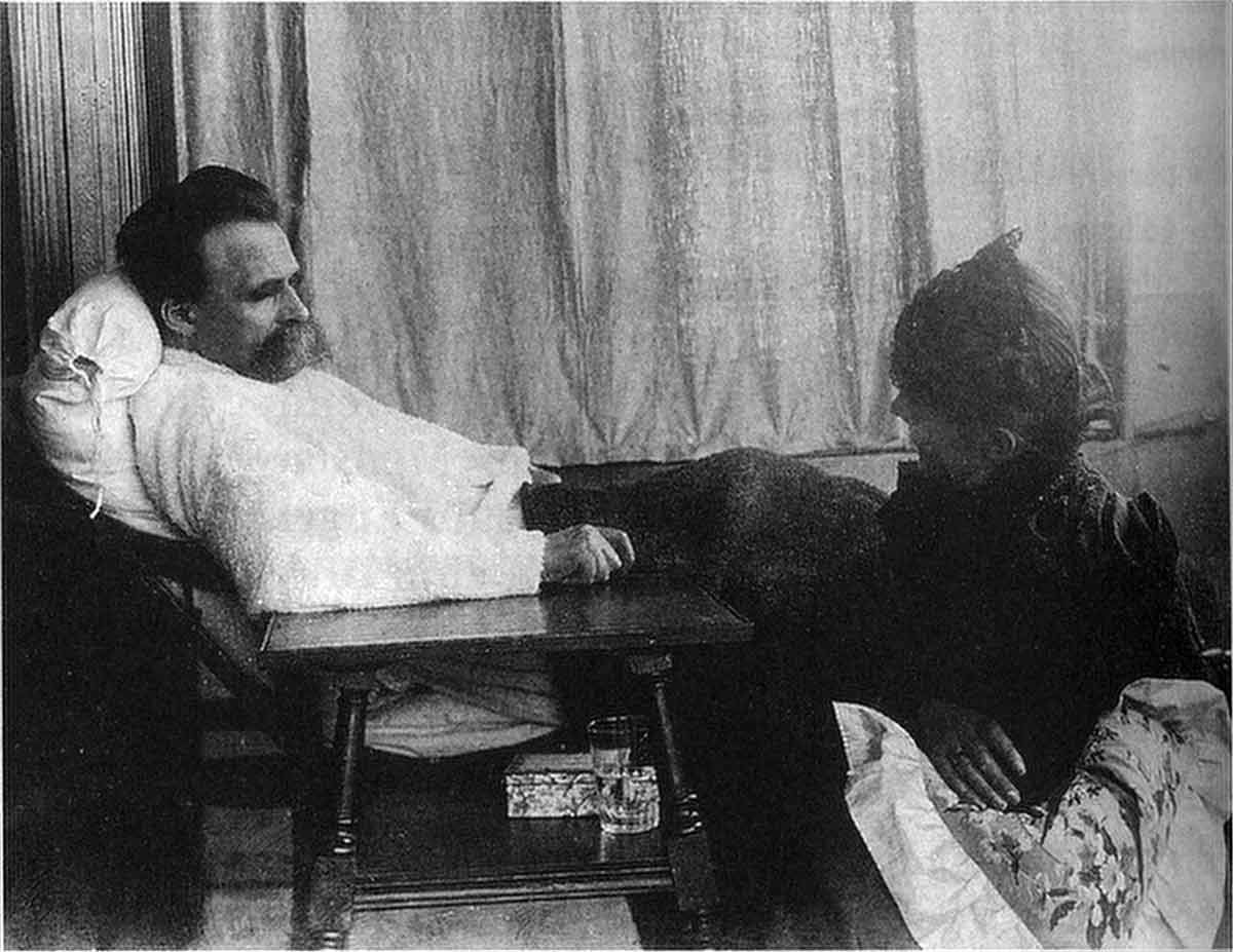 A black and white historical photograph showing an ailing, older Friedrich Nietzsche with his prominent mustache, propped up against a white pillow on a reclining chair or bed. He wears a thick white garment and rests his arm near a small dark wooden table that holds a glass and a small box. A woman in a dark, high-collared dress sits beside him in the foreground, looking toward him.