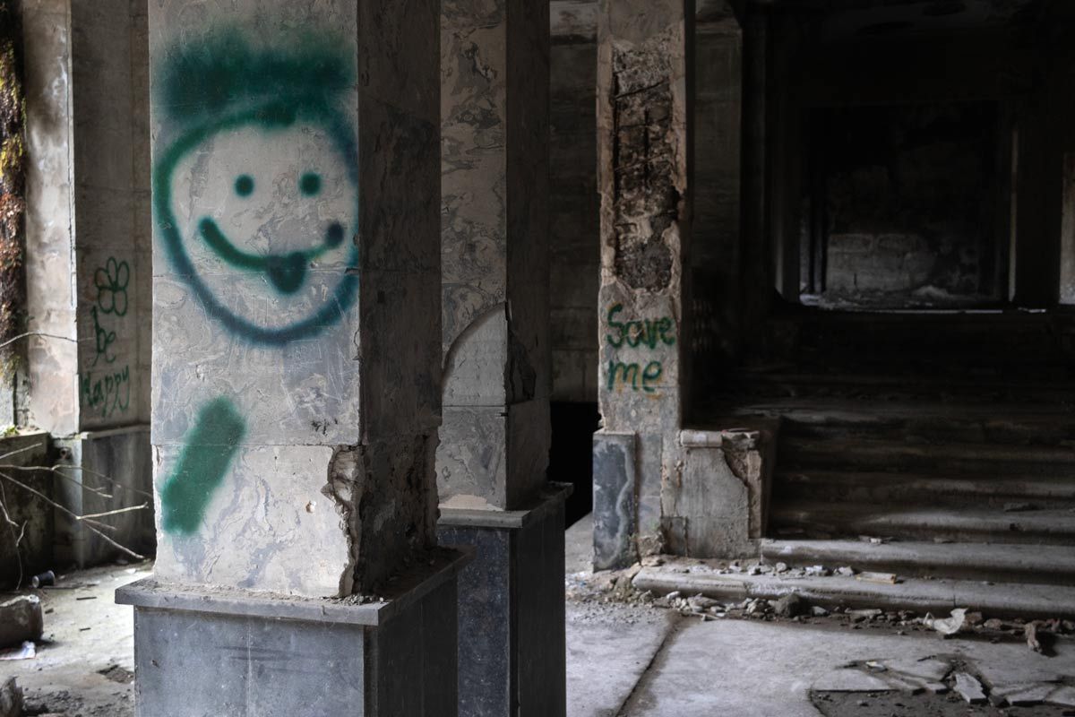 A square stone pillar in a dark, decaying interior featuring a green spray-painted smiley face with its tongue out. In the background, another pillar is marked with the words "Save me" in green paint next to a debris-strewn staircase.