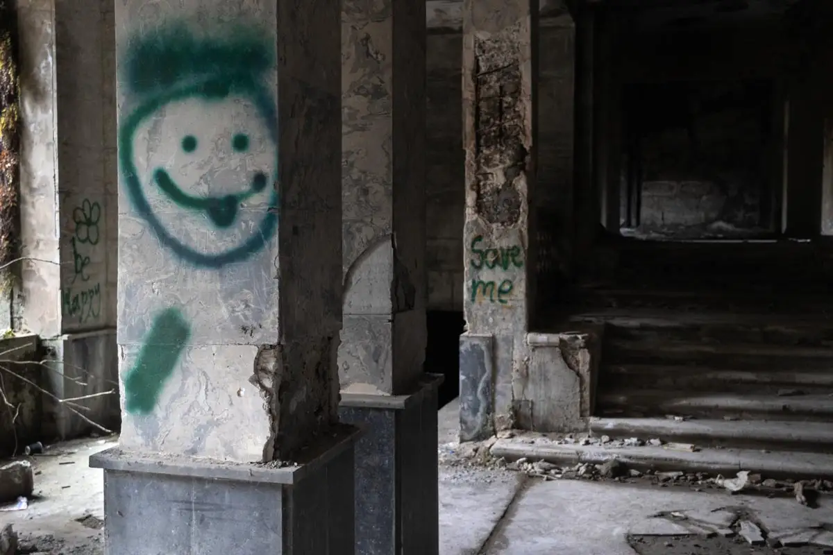 A square stone pillar in a dark, decaying interior featuring a green spray-painted smiley face with its tongue out. In the background, another pillar is marked with the words "Save me" in green paint next to a debris-strewn staircase.