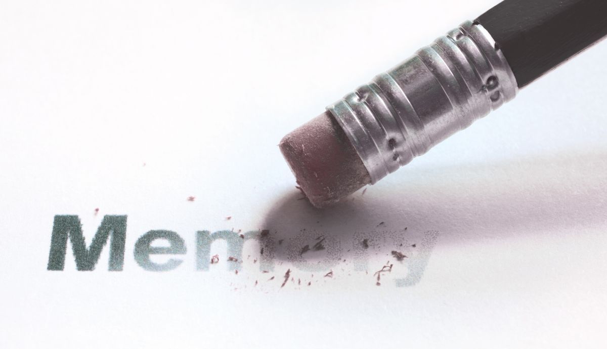 Close-up of a pencil eraser rubbing out the printed word "Memory" on white paper, leaving behind eraser shavings and faded letters.