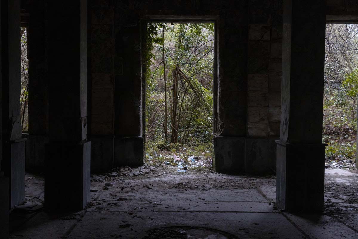 Looking from a dark, shadowed room through a central doorway that is completely blocked by lush, overgrown green vines and trees. Plastic bottles and trash are scattered across the threshold in the foreground.