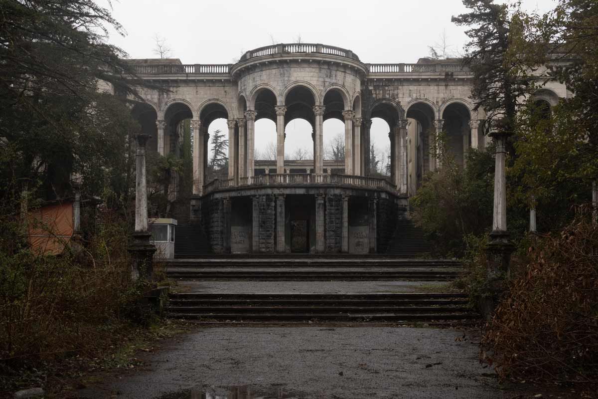A wide shot of the ruined Sanatorium Medea under a grey sky, featuring a symmetrical neoclassical portico with stone arches, a central rotunda, and wide stairs leading into the dark interior.