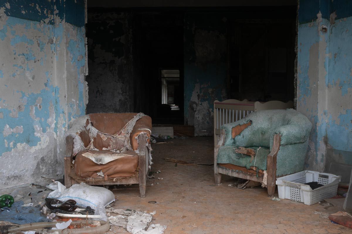Two heavily worn and ripped armchairs—one brown and one teal—sit in a room with peeling blue paint. A white wooden crib is tucked into the shadows of the hallway behind them, surrounded by scattered debris.