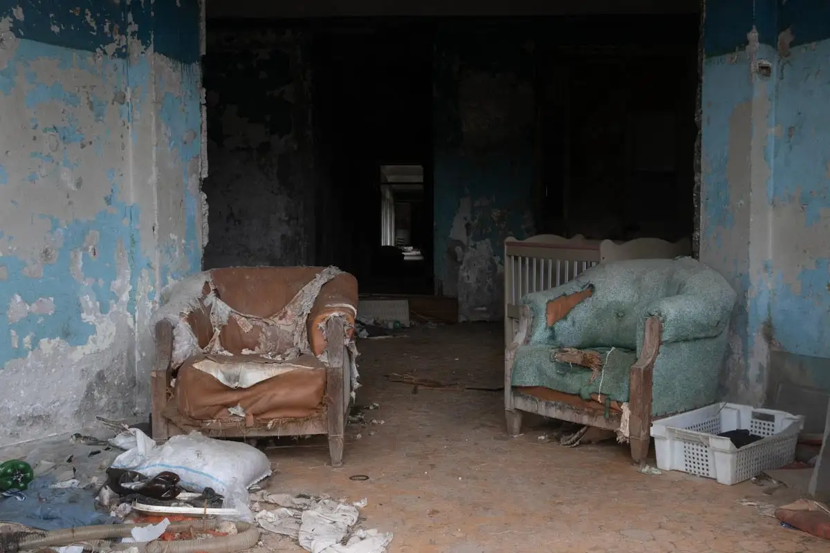 Two heavily worn and ripped armchairs—one brown and one teal—sit in a room with peeling blue paint. A white wooden crib is tucked into the shadows of the hallway behind them, surrounded by scattered debris.