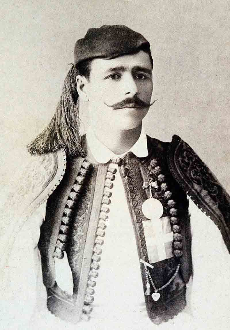 A black and white portrait of a man with a prominent mustache wearing traditional Greek attire, including a tasseled cap and a heavily embroidered vest pinned with medals.   