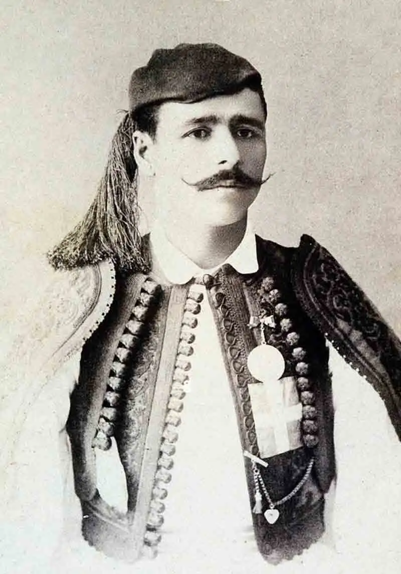 A black and white portrait of a man with a prominent mustache wearing traditional Greek attire, including a tasseled cap and a heavily embroidered vest pinned with medals.