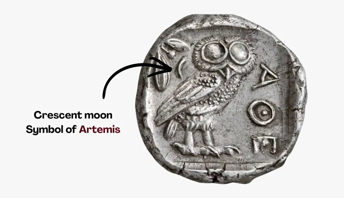 A close-up of an ancient silver Athenian coin featuring an owl on one side, with an arrow pointing to a small crescent moon symbol.