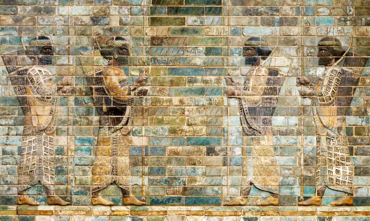 A colorful ancient frieze made of glazed bricks depicting four Persian archers in profile. The soldiers have dark skin and curly black beards, wearing richly patterned robes in shades of yellow, brown, and white. They are shown mid-stride, carrying spears and with bows and quivers slung over their shoulders against a background of blue and teal bricks.