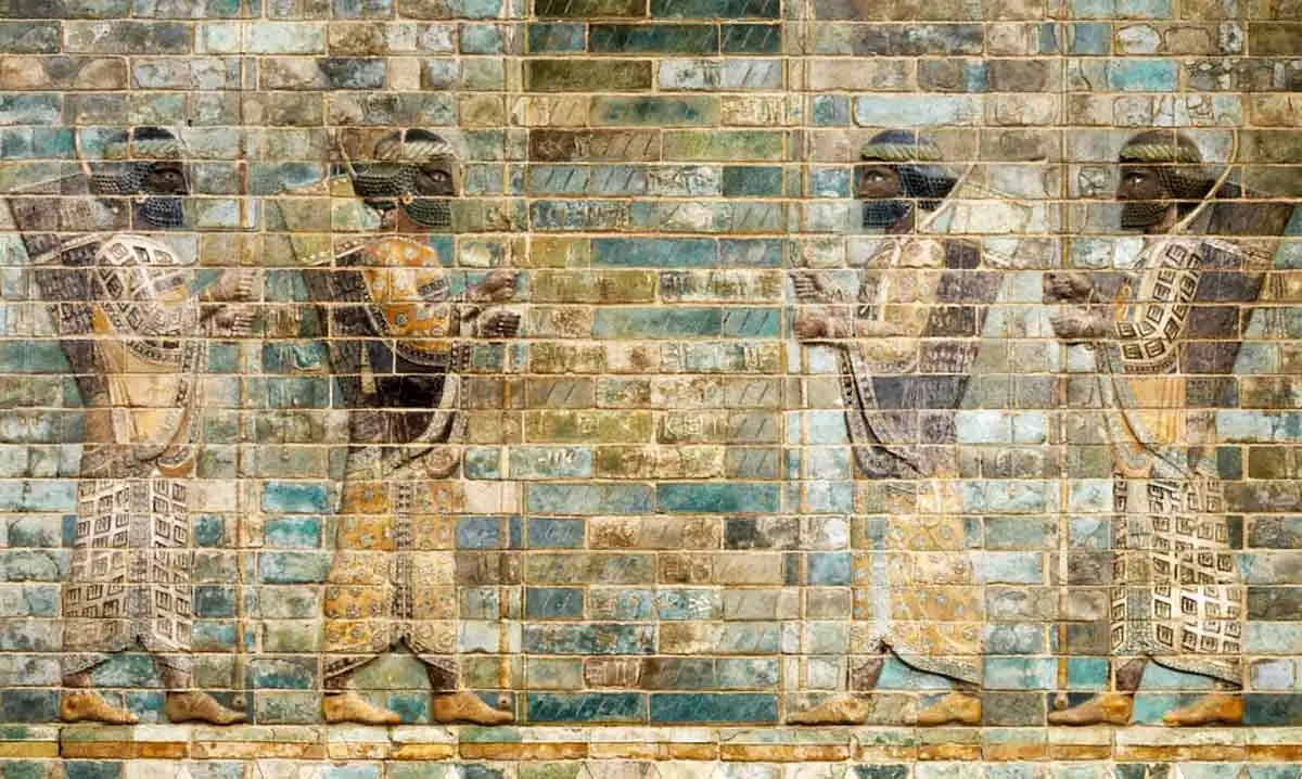 A colorful ancient frieze made of glazed bricks depicting four Persian archers in profile. The soldiers have dark skin and curly black beards, wearing richly patterned robes in shades of yellow, brown, and white. They are shown mid-stride, carrying spears and with bows and quivers slung over their shoulders against a background of blue and teal bricks.
