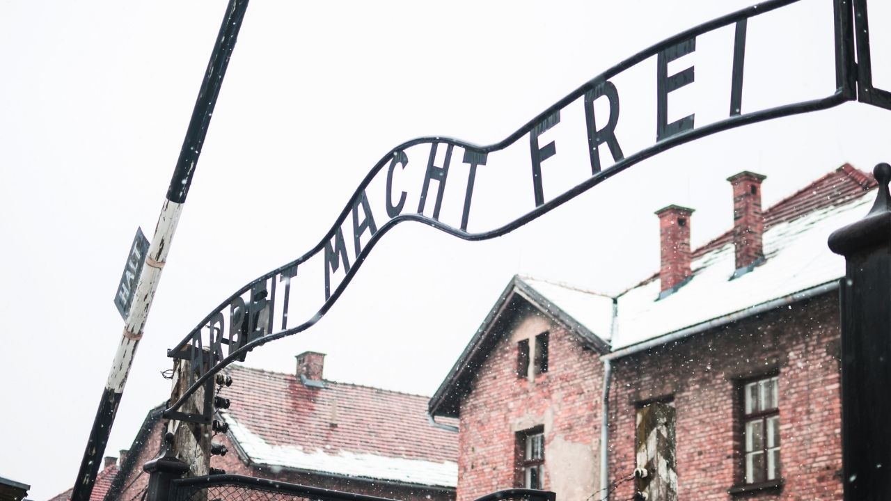 Looking up at the curved metal "ARBEIT MACHT FREI" sign arching over the gate of Auschwitz, with snow falling and brick barracks visible in the background. A partial "HALT" sign is visible on the left post.
