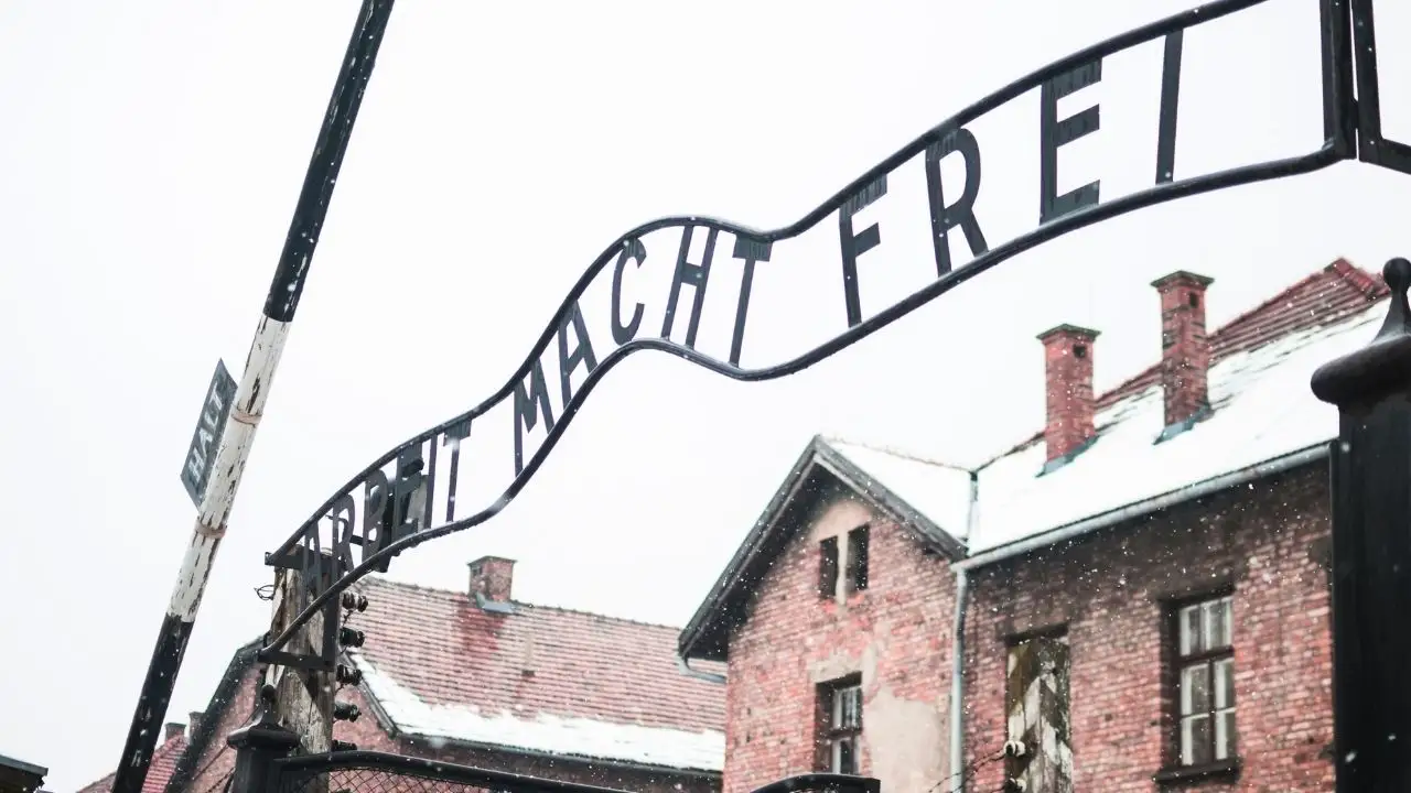 Looking up at the curved metal "ARBEIT MACHT FREI" sign arching over the gate of Auschwitz, with snow falling and brick barracks visible in the background. A partial "HALT" sign is visible on the left post.