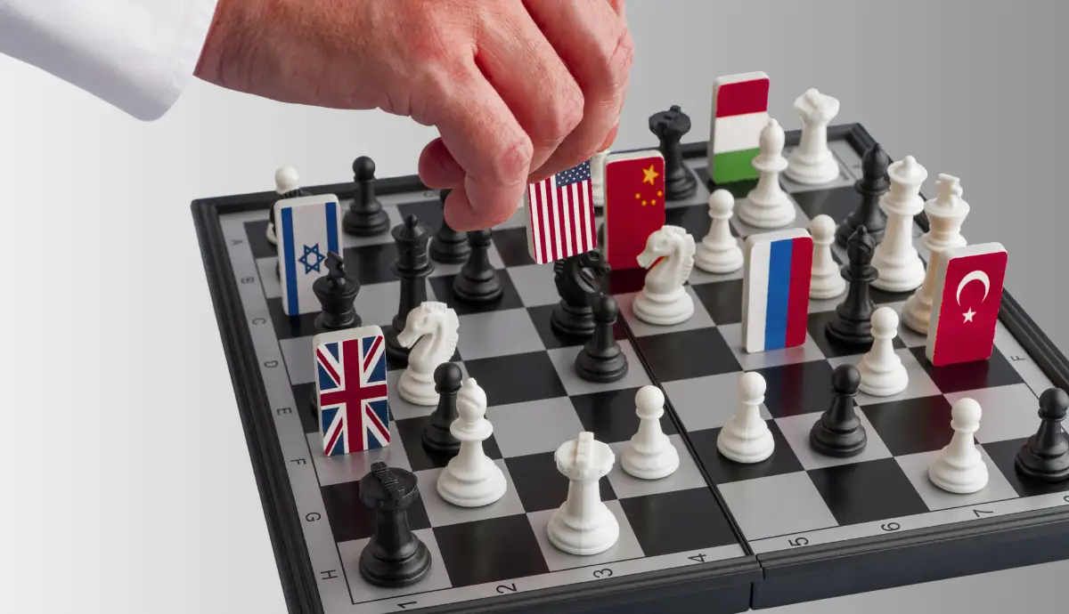 A hand moves a chess piece marked with the United States flag on a chessboard. Several other pieces on the board display national flags, including those of China, the United Kingdom, Russia, Israel, Turkey, and others.