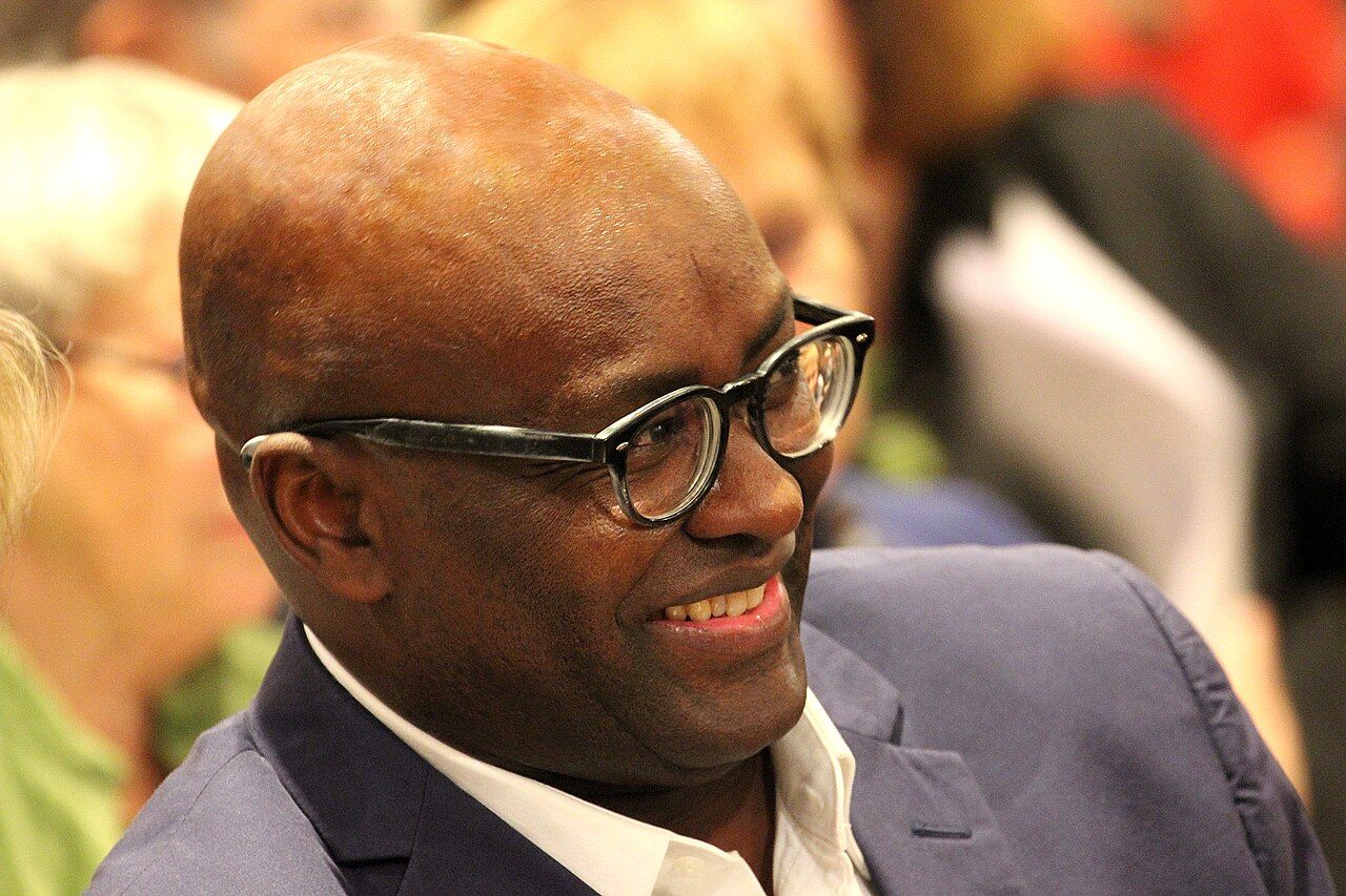 A portrait of the philosopher and historian Achille Mbembe. He is shown from the chest up, wearing glasses and a dark, textured jacket, looking directly at the camera with a neutral expression. The background is a blurred interior with bookshelves.
