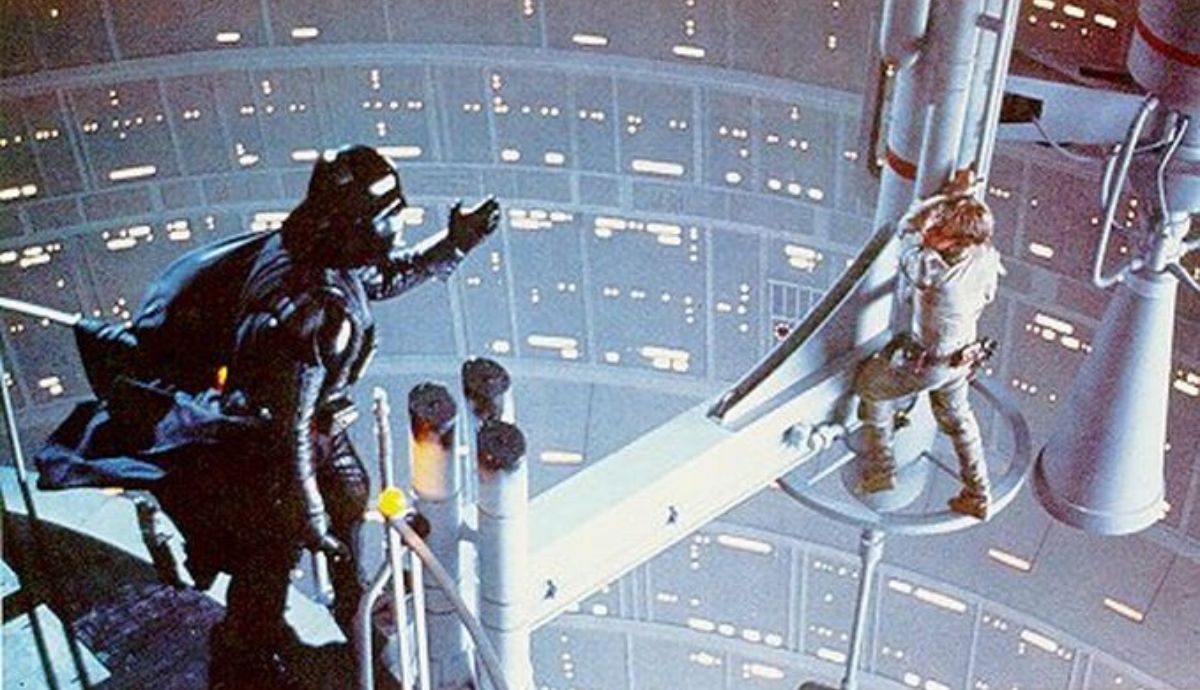 Cinematic still from Star Wars: The Empire Strikes Back showing Darth Vader gesturing toward Luke Skywalker on a narrow gantry inside Cloud City.