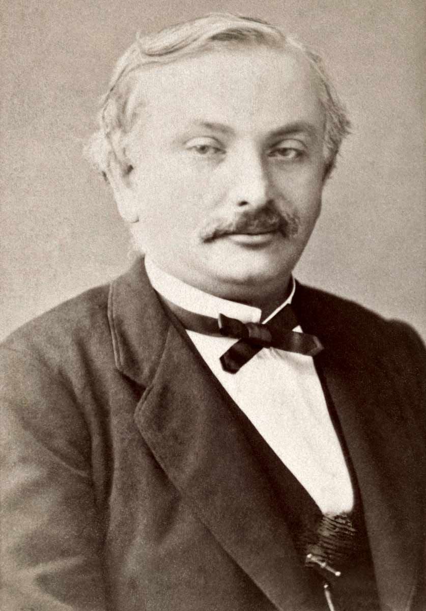 A sepia-toned portrait photograph of a man with a mustache, dressed in a dark suit jacket, vest, white collared shirt, and a dark bow tie.   