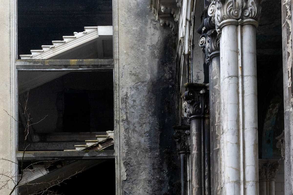 A detailed architectural shot showing a white staircase visible through a concrete window frame. To the right, weathered double columns with ornate Corinthian capitals stand against a dark, textured wall.