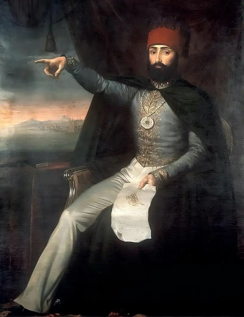 An oil painting of Ottoman Sultan Mahmud II seated on a chair, wearing a red fez and a blue military jacket with gold embroidery while pointing his right arm toward a distant harbor view.