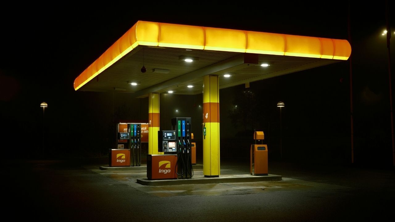A brightly illuminated 'ingo' gas station at night, standing out starkly against the pitch-black surroundings. The station features a modern yellow and orange canopy over several fuel pumps, appearing completely empty of any people or vehicles.