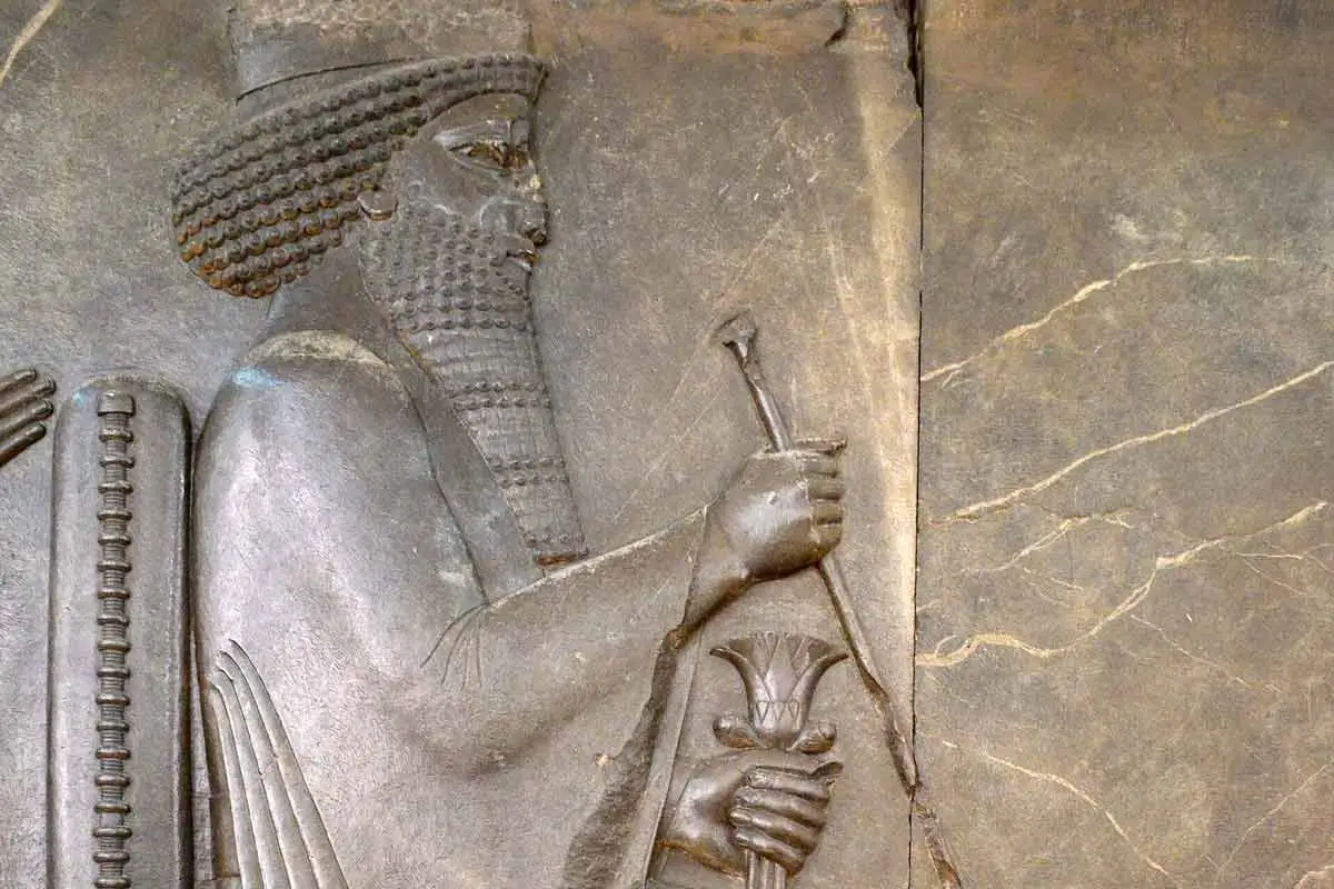 A side-profile stone relief sculpture of a Persian king, likely Xerxes, seated on an ornate throne. He wears a long, draped robe, a crown, and has a meticulously groomed, long beard. He holds a long scepter in his right hand and a lotus flower in his left. The stone shows signs of age with several cracks across the surface.