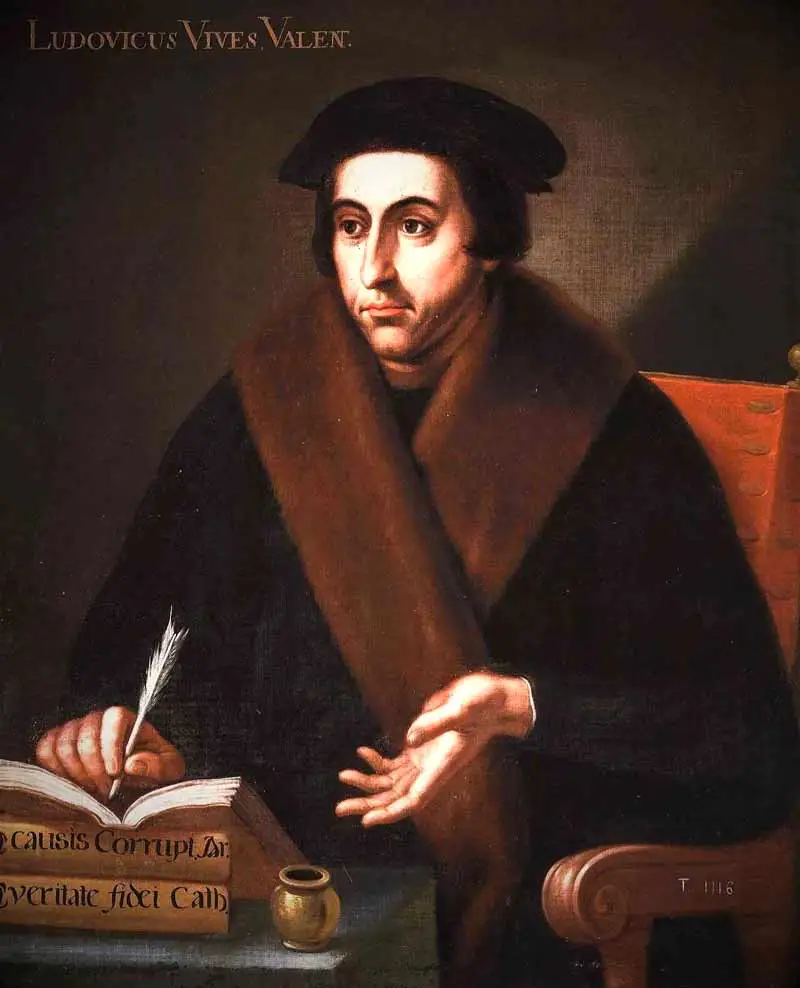 A historical oil painting of Juan Luis Vives seated in a wooden chair at a desk. He wears a black cap and a dark coat with a thick, brown fur collar. His right hand holds a quill pen poised over a thick, open book, while his left hand rests open in a gentle upward gesture. A small gold inkwell sits beside the book on the table. The inscription "LUDOVICUS VIVES VALEN." is painted in the upper left corner against a dark, shadowy background.
