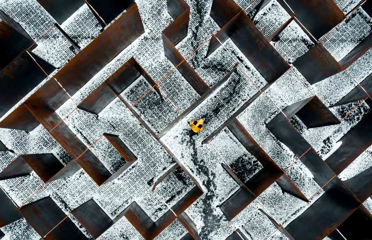 high-angle aerial shot of a complex maze with tall, rusted metal walls. A single person wearing a bright yellow jacket stands in the center of one of the snowy pathways, creating a sharp contrast against the industrial browns and grays. The ground of the labyrinth is covered in a mix of light snow and dark, slushy footprints that trace a path through the corridors.