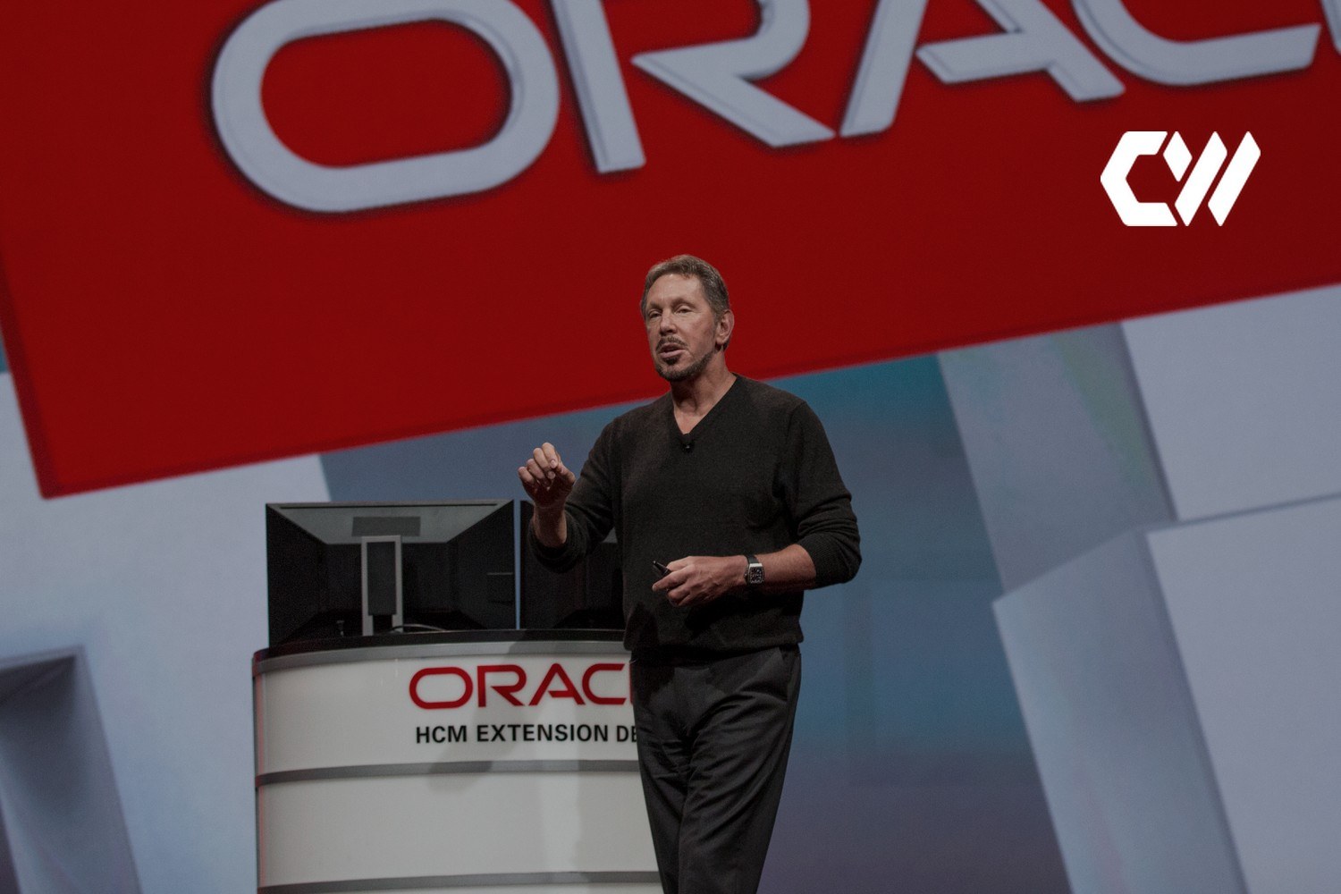 Larry Ellison presenterer på en Oracle-scene med en stor Oracle-logo i bakgrunnen. Med logoen til CoreWeave lagt på.