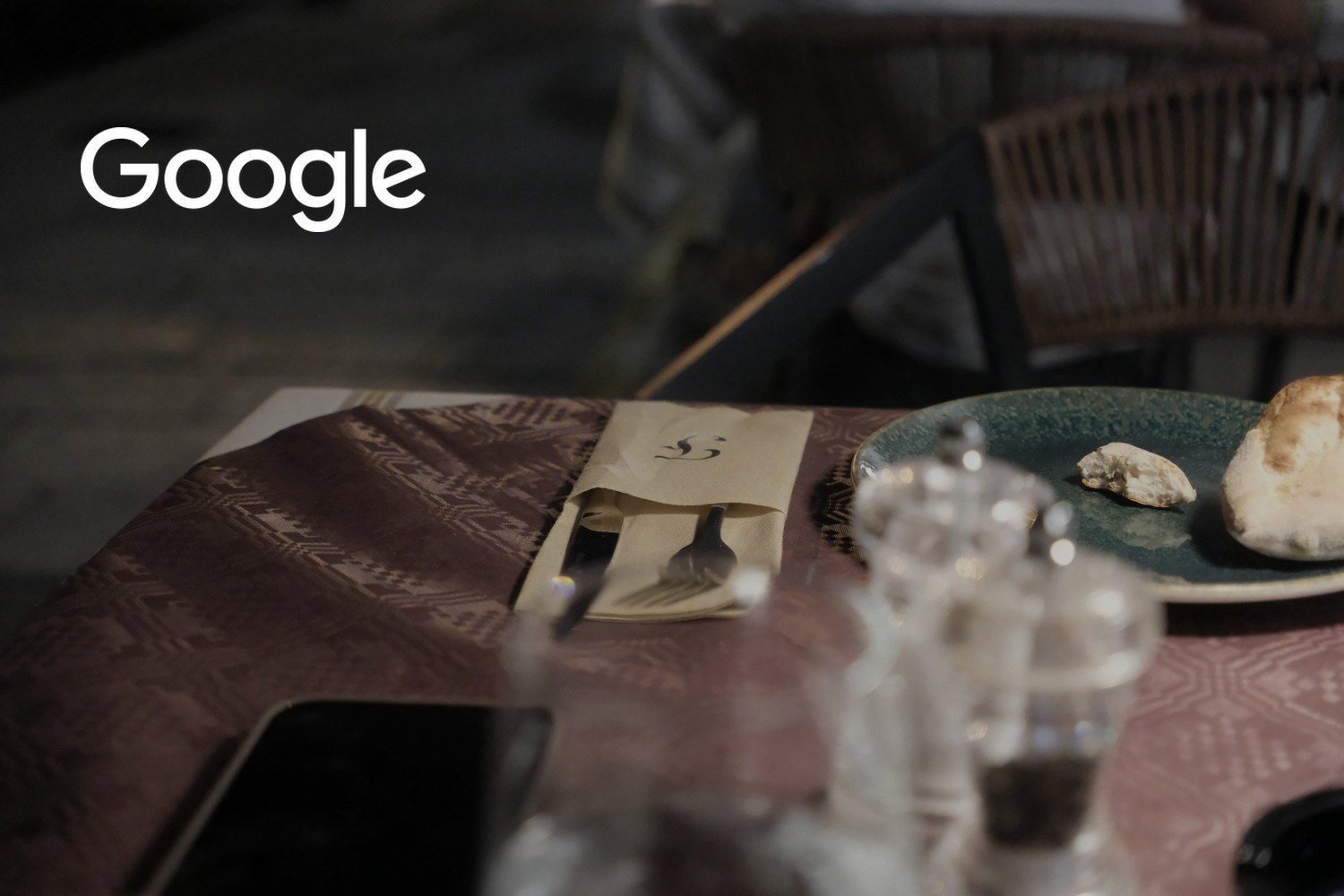 En mobiltelefon ligger på et dekket restaurantbord med bestikk, brød og tallerken. Med logoen til Google lagt på.